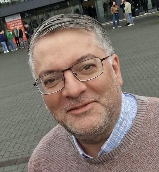 Mann mit Brille und grauen Haaren, trägt einen braunen Pullover, lächelt freundlich.